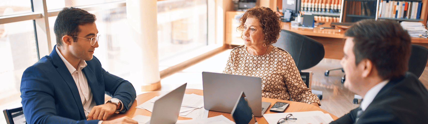 Three business colleagues gathered around discussing the concept of intelligence elevated with their opened laptops and data spreadsheets on top of a wooden table in a well-lit office room