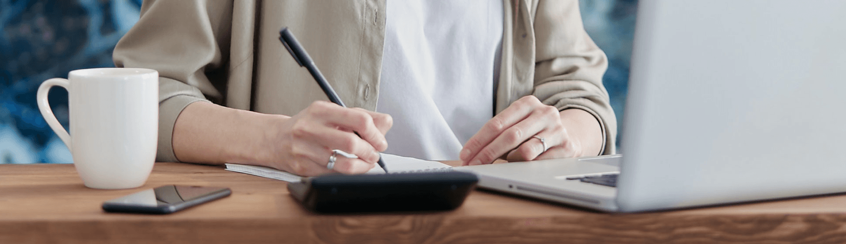 A focused shot of a woman taking notes on her desk with an opened laptop, a ceramic mug, and a calculator being displayed
