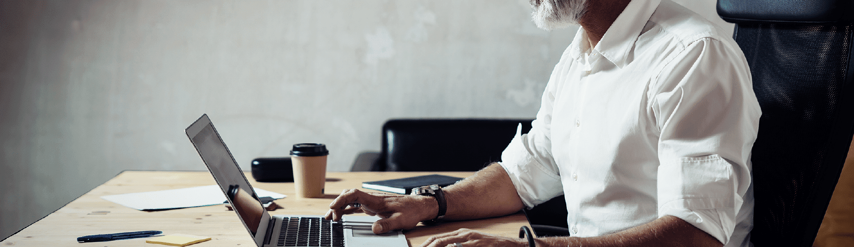 Older Data Analyst gentleman sitting on his desk with an opened laptop, coffee cup, and notebook