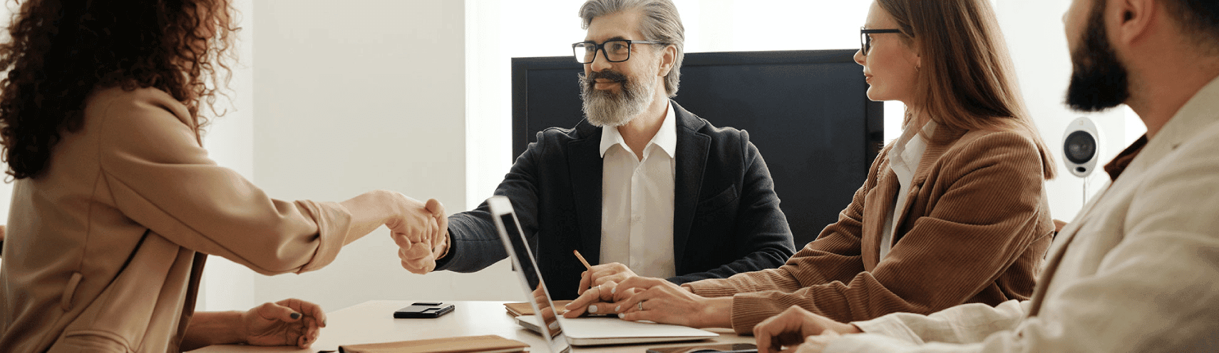 A group of four business people sitting around a desk with their laptops as one of them shakes another's hand